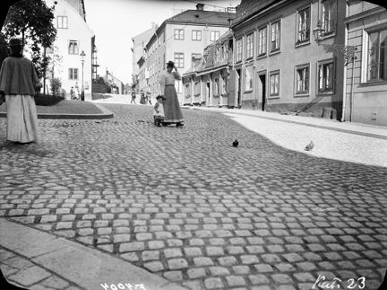 Flera personer syns i bild, gående över den gatstensbelagda backen. Mitt i bild går en kvinna och ett litet barn hand i hand.