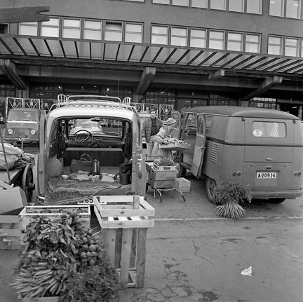 En kvinna och en man lastar in frukt och gr&#xF6;nsaker i en sk&#xE5;pbil. Intill st&#xE5;r en annan sk&#xE5;pbil med bakd&#xF6;rrarna &#xF6;ppna. Tr&#xE4;l&#xE5;dor med stora knippen av mor&#xF6;tter st&#xE5;r p&#xE5; marken.