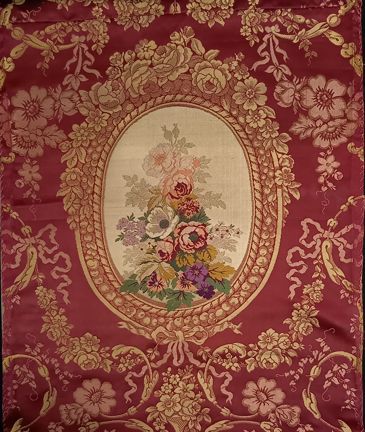 Ett rött tyg med gula och rosa blomstergirlanger runt en oval ram. I ramens mitt visas en färggrann blombukett i många färger som grönt, lila, gult, grönt, rött, violett, rosa mot vit bakgrund.