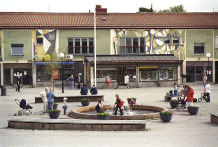 Vuxna och barn rör sig på torget. I mitten en fontän.