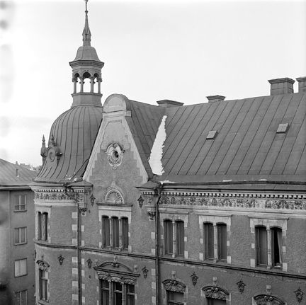 Tak och översta våningsplanet av ett högt flerfamiljshus av sten. Ett torn pryder det rundade hörnet. Tornet har överst en liten lanternin. Fasaden har dekorativa utsmyckningar.