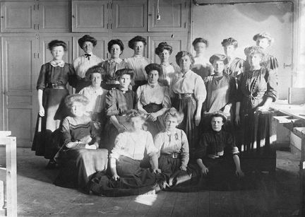 En gruppbild med arton kvinnor. Några står, några sitter, framför en vägg med skåp. Ljuset faller in från ett fönster till höger i bild. Alla kvinnorna bär långkjol och har håret uppsatt i valk och knut.