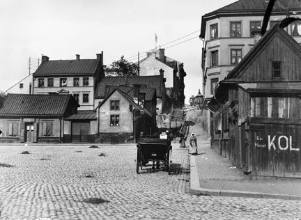 Runt ett torg belagt med gatsten, ligger flera små hus av sten och trä. Intill trottoaren står en häst med vagn. Två kvinnor kommer gående, annars är det i stort sett folktomt. Uppe på en höjd ligger några något större stenhus. På ett plank står det "KOL".