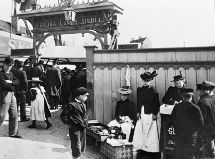 Vid entrén till ett tivoli flockas några pojkar kring de kvinnliga försäljarna som står utanför. Det ser ut att säljas glaserade munkar och andra godsaker. Flickorna har ett högt träplank bakom sig och till vänster om det syns en hög portal av snidat trä. Många människor är i rörelse och på väg in genom entrén.