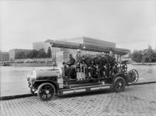 Brandbil, troligen en Scania Vabis, med brandmän  på Södra Blasieholmskajen.  I bakgrunden Slottet. Troligen är det brandkårens första motorspruta som fotograferas 30 juli 1918.