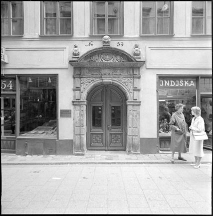 Till vänster om portalen en butik med tavlor liggande i skyltfönster, till höger en Indiska-butik., där två kvinnor står och pratar på trottoaren nära skyltfönstret.