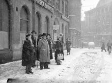 Gatumiljö i snöväder. Folksamling vid hållplats