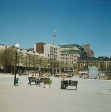 Öppen plats med parkbänkar i Kungsträdgården. Vy åt nordväst mot Sverigehuset, Nordiska Kompaniet och Sagerska husen