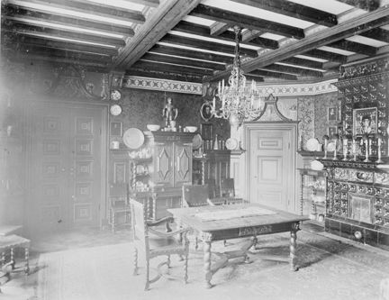 Rum med bjälktak och många utsmyckningar i dörrar, möblemang och kakelugn.