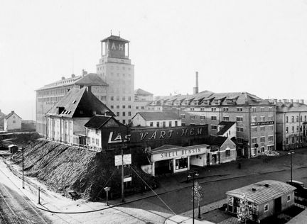 På en liten bergknalle intill en större gatukorsning, ligger några små och ålderdomliga hus kvar. Bakom dem reser sig en stor och nybyggd varuhusbyggnad med torn. Intill ligger en kontorsbyggnad och några bostadshus. I bildens högra hörn ligger en låg kioskbyggnad.