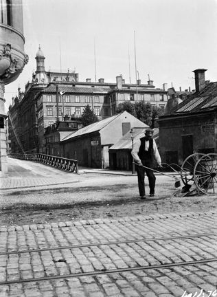 En man iförd keps, vit skjorta och väst sopar en gata. Bredvid står en skottkärra av äldre modell med stora hjul. I förgrunden syns gatstenar med spårvagnsspår. Nära mannen syns ett ornerat burspråk. I bakgrunden lägre stenhus och bortom dem högre stenhus. Gatan bakom mannen löper i två nivåer och avskiljs med ett lågt staket.