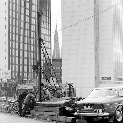 Pålningsarbete i kv. Putten. Klara kyrkas torn syns mellan fjärde och femte hötorgshusen. Tre män arbetar runt pålningsmaskinen. I förgrunden syns en bil.