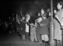 Kung Gustaf V:s kista förs från Drottningholms slott till slottskapellet i Kungliga slottet i Stockholm. Utanför slottet har folk samlats med facklor i händerna