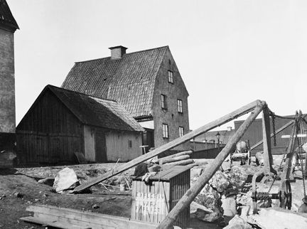 En man, knappt synbar, står med ryggen vänd mot kameran, mitt i en byggarbetsplats. Runt omkring honom finns stora stenblock och över honom en byggnadsställning. I bakgrunden syns en liten träbod och ett större putsat hus med sadeltak.