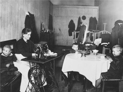 Bilden visar en kvinna som sitter och syr vid en symaskin och tre barn som sitter vid bordet.