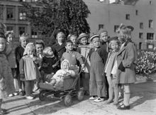 Grupporträtt av en samling barn vars mammor vid tillfället ägnar sig åt husmodersgymnastik. Här passas de av barnträdgårdslärarinnan Margot Fränckels