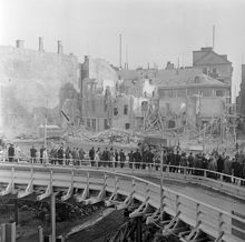Folksamling på provisoriska träbron vid Mäster Samuelsgatan 45. Vy söderut med rivning av kvarteren Stigbygeln och Hoven. Numera platsen för 5:e höghuset. I bildens högra kant går Beridarbansgatan.