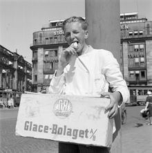 Tegelbacken. En ung glassförsäljare äter glass, med en glasslåda från Glace-Bolaget (GB) på magen. Centralpalatset i bakgrunden