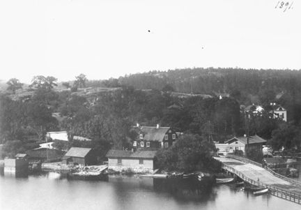 Liljeholmsbron till höger i bild, äldre bebyggelse ligger i grönska. Några småbåtar ligger förtöjda vid bron och bryggor.