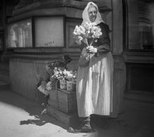 Hötorget. Äldre kvinna klädd i sjalett och förkläde säljer blommor från en korg.