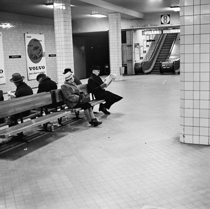 En tunnelbanestation under jord där väggar och pelare är klädda med ljust kakel. På bänkar sitter väntande passagerare, bland annat en man som läser tidningen. I fonden en rulltrappa. På väggarna syns stora reklamtavlor.