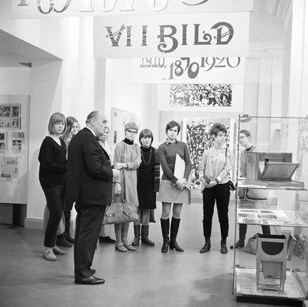 Inifrån en musei-lokal med skylt i taket som visar utställningens titel, Vi i bild. Under skylten står en grupp på sju unga kvinnor samlade och lyssnar till en visning som hålls av en äldre man. Till höger syns en glasmonter med foto-föremål.