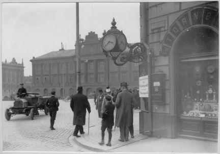 En bil körs över torget och människor rör sig utanför uraffären. En stor klocka sticker ut från byggnadens hörn och i skyltfönstret  syns en mängd klockor.