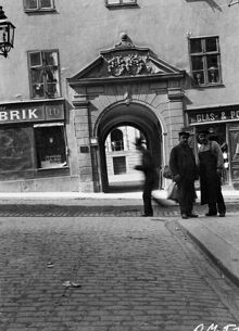 Några män står i hörnet av Mariagränd och Götgatan. I bakgrunden ligger portalen till Götgatan 16, Ebba Brahes palats
