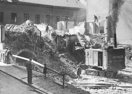 Ett hus rivs med hjälp av en grävskopa. Det ryker och dammar. Ett källarvalv syns till vänster. En man och en pojke tittar på. 