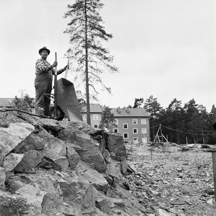 Man med skottkärra på en bergknalle. Hus i bakgrunden.