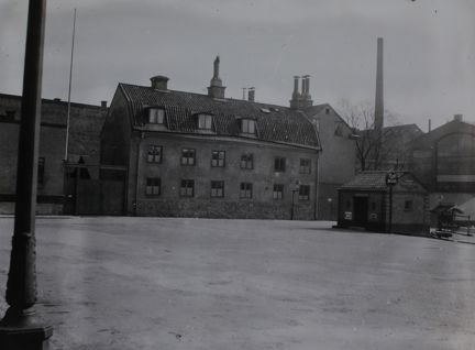 Vid ett ödsligt torg ligger ett väldigt litet hus som ser ut att vara en sorts tank- eller pumpstation. I bakgrunden syns ett par trevåningshus av sten.