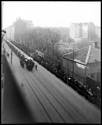 Vy över författaren August Strindbergs begravningståg på väg norrut längs Norrtullsgatan mot Norra Begravningsplatsen. Till höger byggnaden vid Norrtullsgatan 67 i kvarteret Rundellen.