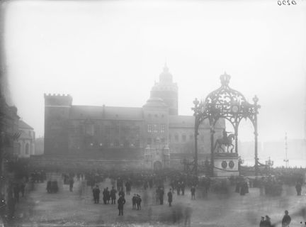 Dimman ligger tät över ett torg. Mitt på torget står det en staty av en ryttare till häst. Statyn har klätts in i en krokanliknande dekoration. I bakgrunden syns en stor kuliss som föreställer en äldre slottsbyggnad. På torget har många människor samlats.