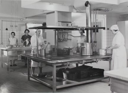 Fem kvinnor i ett storkök. Tre av kvinnorna står runt en stor metallspis med stora kastruller. Kvinnorna har vita rockar och vita huvuddukar.