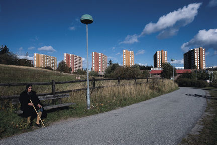 Kvinna med stav sitter på parkbänk längs gångväg.