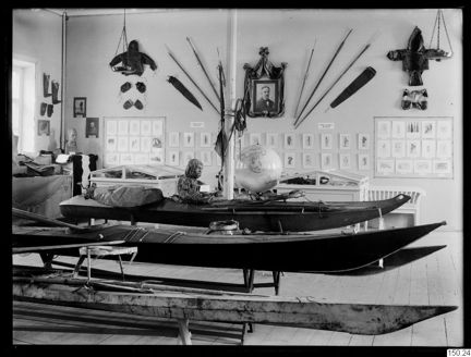 Svartvitt fotografi som visar ett ljust utställningsrum med kajaker och andra föremål.