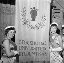 Hantverkargatan 1. Stockholms högskola blev Stockholms universitet. Invigning av det nya universitetet firas i Stadshuset. Två flickor i studentmössa och långklänningar står vid studentkårens fana