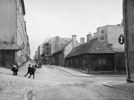 En gatuvy med stenläggning. Några barn står i gathörnet och tittar mot fotografen. Till höger syns gammal trähusbebyggelse, längre bort är byggnadsställnigar resta. Längst fram i högra hörnet syns en skylt
