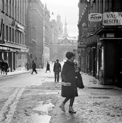 Kvinna med paket i handen i förgrunden. Butiksskyltar.