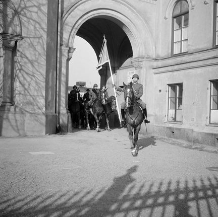 Hästar med ryttare kommer ut på en gårdsplan genom ett valv.