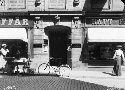 framför en port står en cykel parkerad. Till höger ligger en hattaffär. Två kvinnor i sommarhattar och långa kjolar går på trottoaren. 