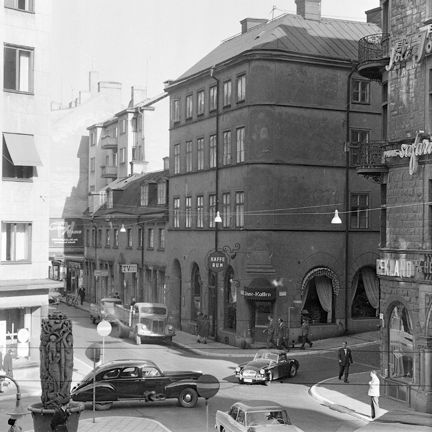 I en vägkorsning ligger ett äldre hus som på hörnet har en ingång till ett kafé. Kaféet har ett dekorativt tak över ingången och stora fönster ut mot gatan. Flera bilar av lite dyrare märken rullar förbi.