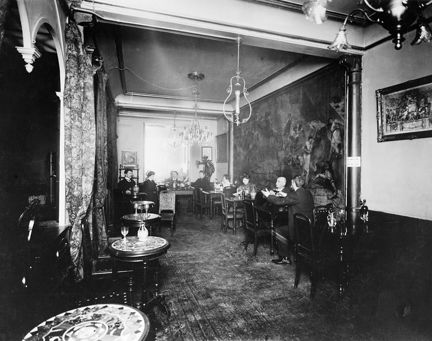 Ett långsmalt restaurangrum med trägolv, träpanel längs väggen och en större väggmålning. Vid små bord med plats för tre personer sitter 1-2 personer, mest män. Kristallkronor och lampetter hänger i taket. 