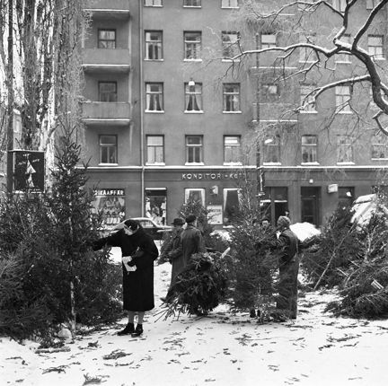 Några kunder och försäljare bland granarna. Snö på marken. I bakgrunden skymtar en trikåaffär och ett konditori.
