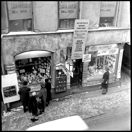 Några människor står framför en butik och tittar i skyltfönstren. Stora skyltar talar om att det är realisation eftersom huset skall rivas. 