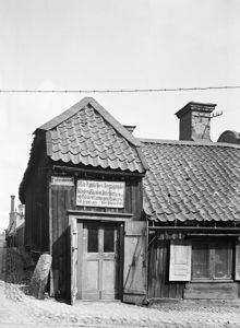 Hörnhuset Lilla Badstugatan / Kungstensgatan 30. På skylten ovanför dörren står det: Här uppköpes begagnade kläder, skodon, buteljer, alla slags metallskrot, lump gummi & diverse till högsta pris