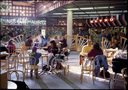 Inomhus i ett köpcentrum. Människor sitter och fikar vid cafébord, i bakgrunden syns en klädbutik och en mataffär..