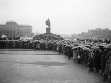 Folksamling vid Slottsbacken på Skeppsbron i väntan på begravningståget med stoftet efter de förolyckade från Andréexpeditionen. I fonden Nationalmuseet och Gustaf III:s staty.