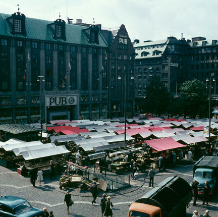 Färgbild som visar ett stort torg med en stor mängd torgstånd. Intill ligger en större varuhusbyggnad med texten "PUB" och "Det finns hos PUB" på fasaden.