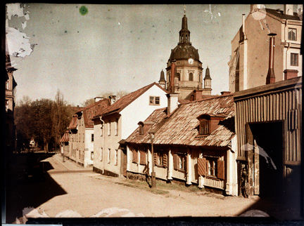 Längs en grusbelagd gata ligger flera ålderdomliga hus i två till tre våningar. En del är av sten, de närmast i bild är av trä. I fonden syns en större kyrkobyggnad med kupolformat tak med ett infällt ur. 